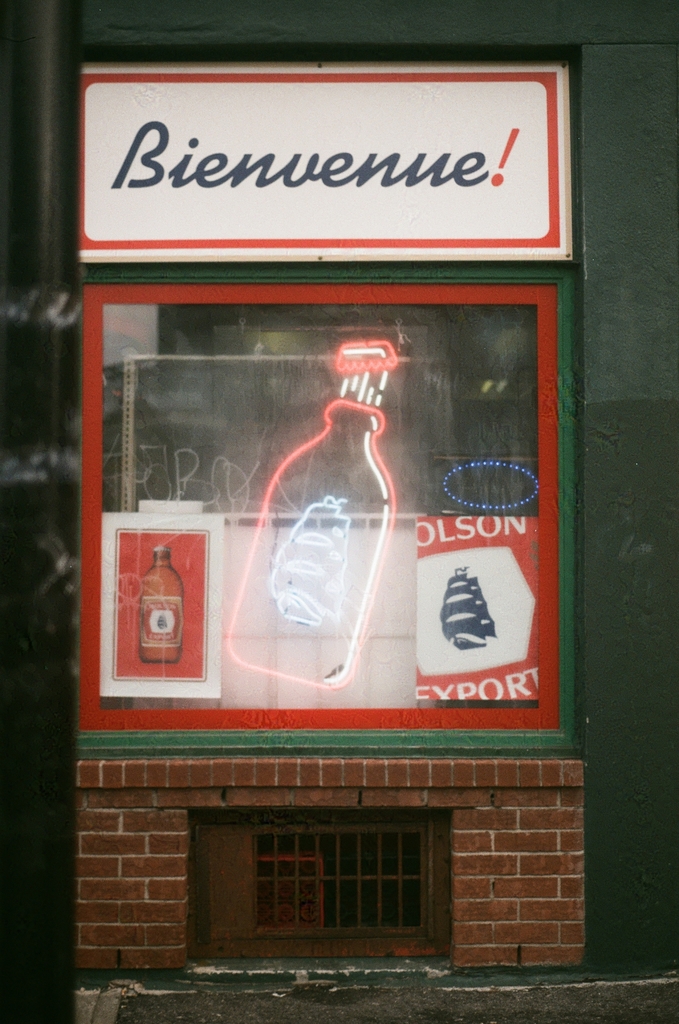 the window of a dépanneur, showing a bottle of Molson Ex in neon, and topped with a big welcome sign reading "Bienvenue!"