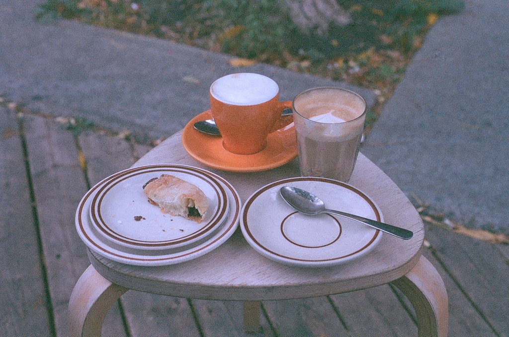 An orange mug of cappuccino & a latte on a stool outside a coffee shop