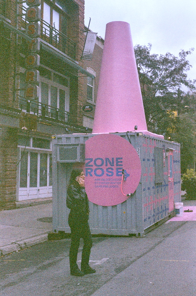 A shipping container with a sign reading Zone Rose, and a giant pink cone rising from the top; a woman stands in front, dressed in black, speaking on a pink phone