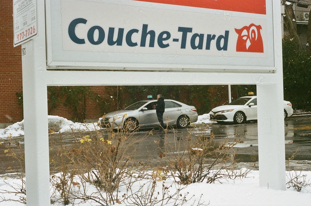 Two parked taxis, one driver standing by the other car & chatting, framed by a Couche-Tard billboard