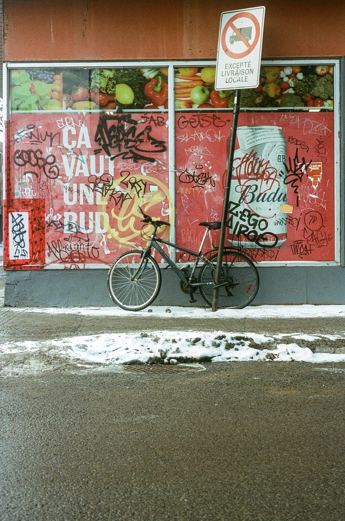 A bicycle is chained to a street sign outside a dépanneur window covered in graffiti
