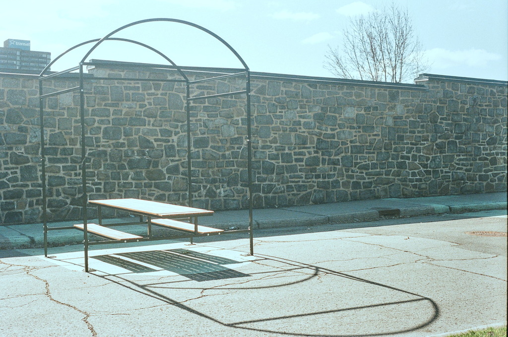A picnic bench on a pedestrian street, with an empty shelter cage around it, casting a long shadow