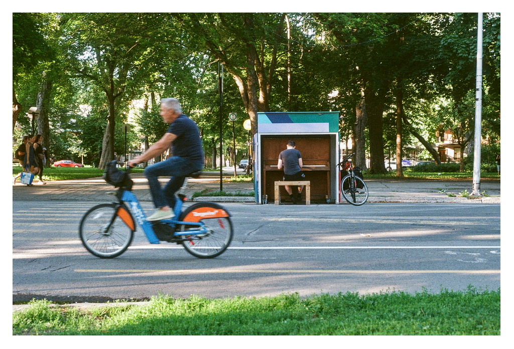 A person is sitting playing a public piano while a trio of pedestrians passes by; a man is blurring by in an electric Bixi in the foreground