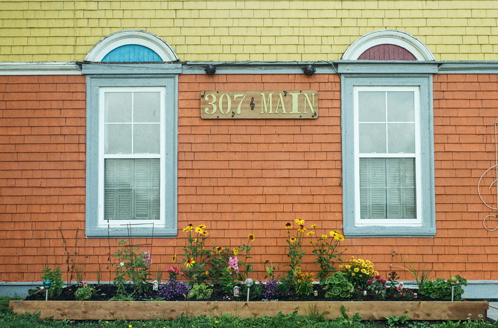 Front of a house, a façade of yellow and orange wooden slats, a flowerbed out front, and the address