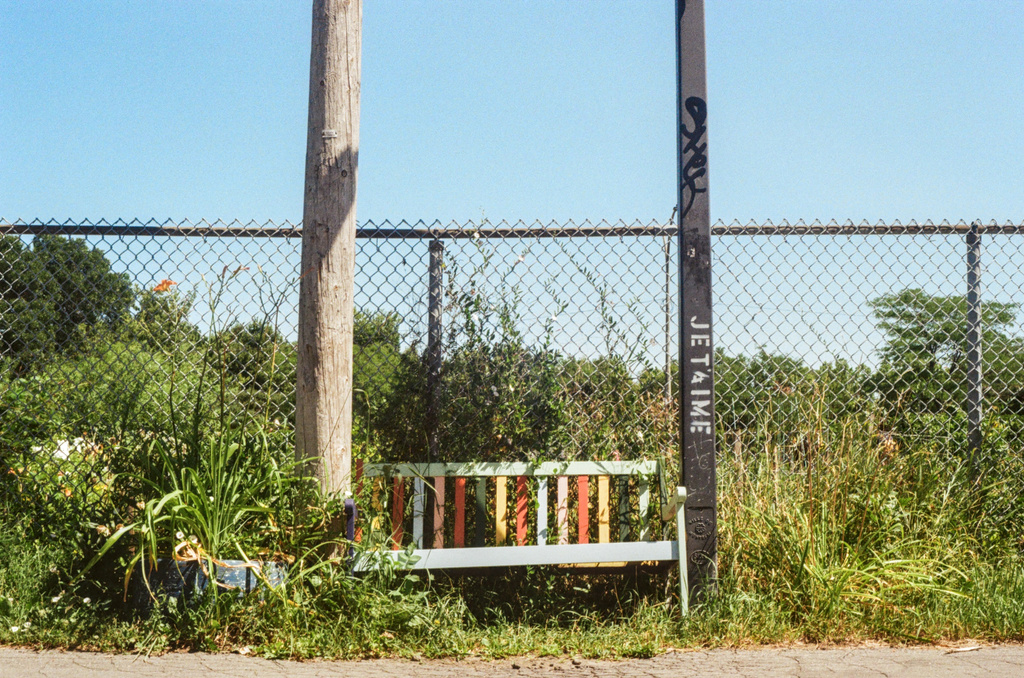 A rainbow-painted bench between a wood and a metal pole, the latter reading "Je t'aime"