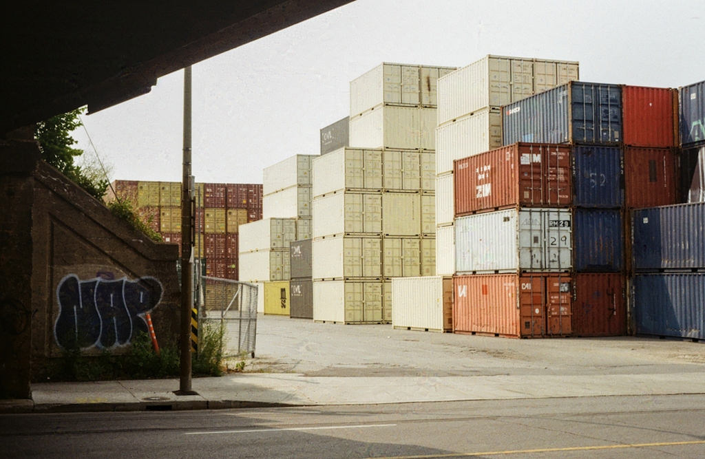Stacks of shipping containers sitting in the sunshine, viewed from under a bridge