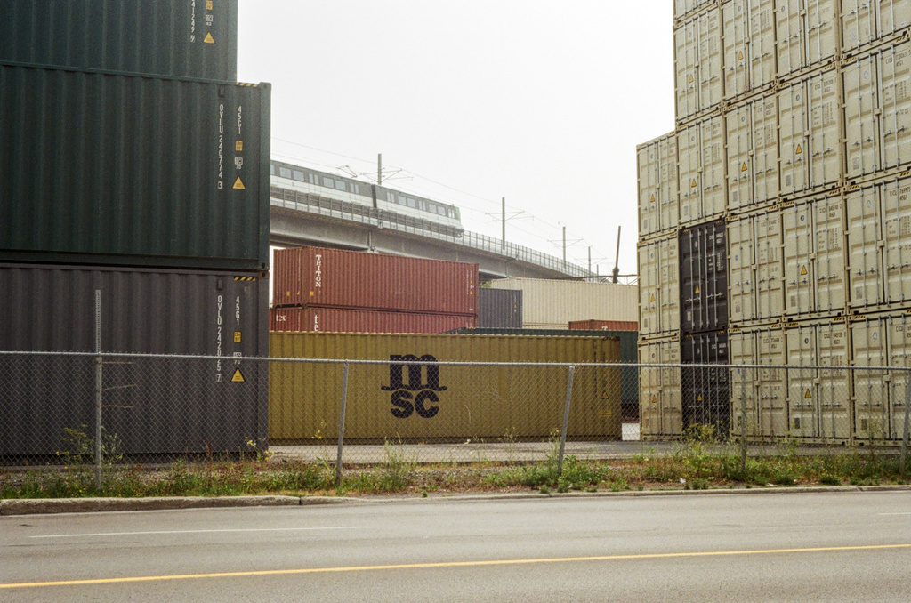 A view through stacks of shipping containers, showing Montréal's light rail REM passing in the background