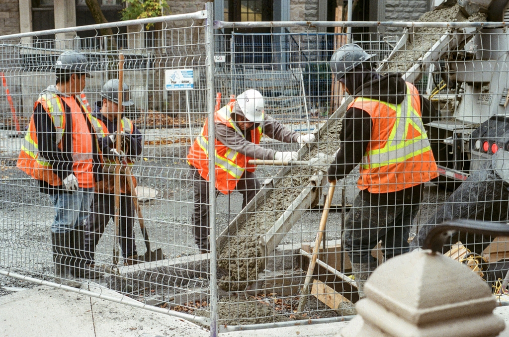 Four workers in high-vis vests pour cement behind a construction fence