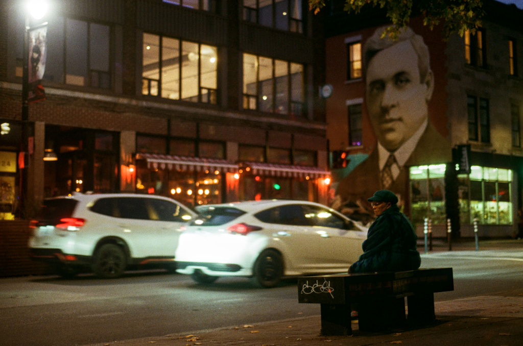 Mural of Aristides overlooking St.-Laurent at night; a woman sits on a bench in the foreground.