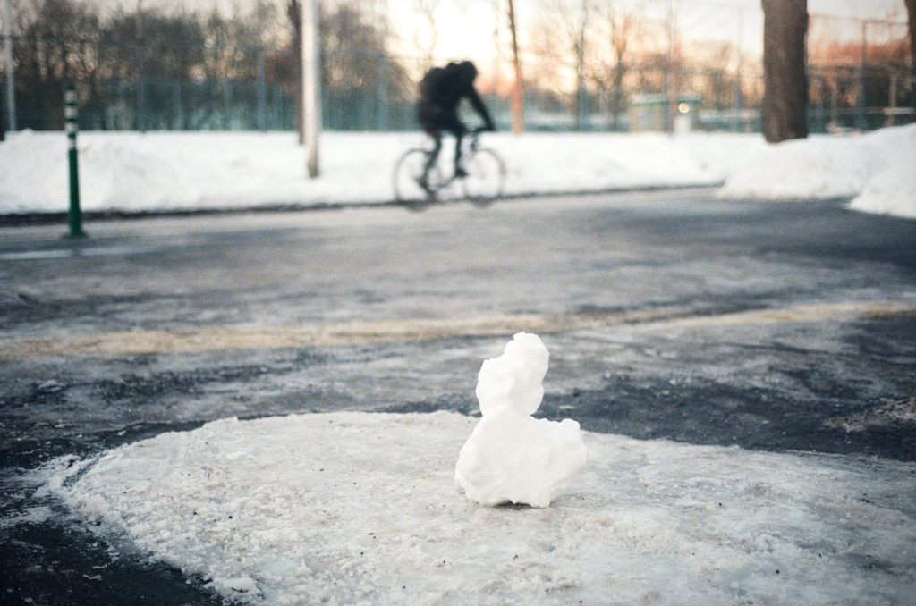 A little snow sculpture slightly resembling a duck; out of focus in the background, a cyclist speeds by