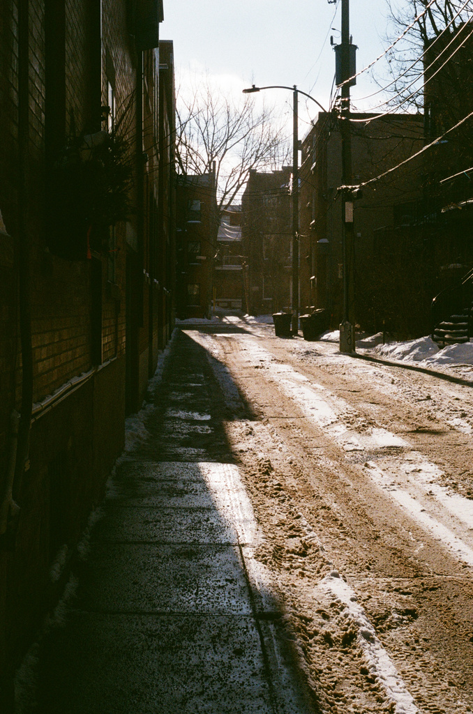 A side street dirty with trampled snow, harshly illuminated by the sun around the corner