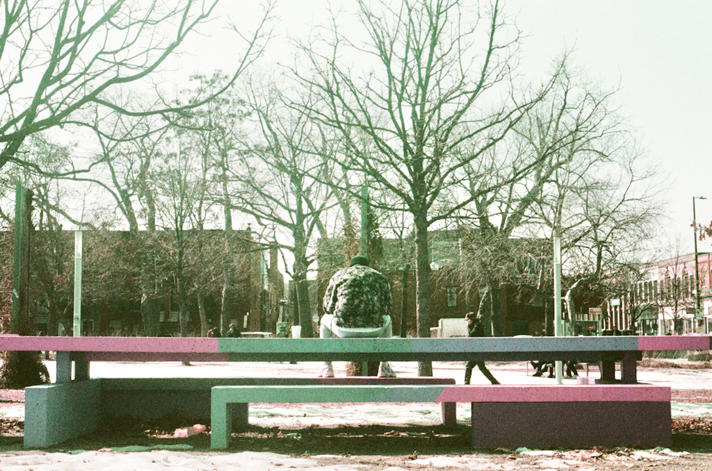 Someone sits on a long park bench or table, facing away from the camera