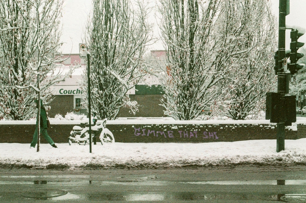 A street corner with a low brick wall, on which someone has spray-painted "Gimme that shi" [sic]. The G has Shrek ears.