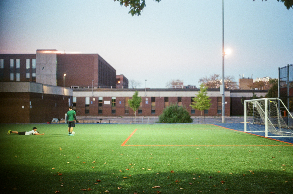 An artificial football field, one person laying on their front, another lining up a shot on net
