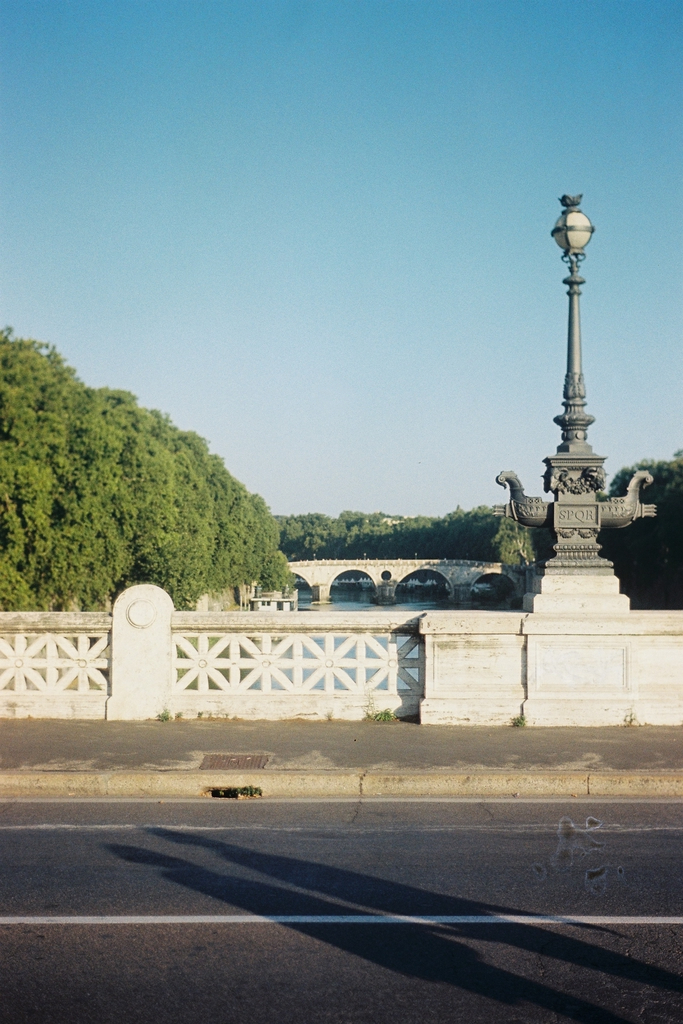 A lamp post, inscribed with SPQR, on a bridge overlooking the Tiber, with two people's elongated shadows